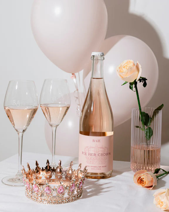 Bottle of pink champagne, two glasses, a decorative tiara, and balloons on a white surface.